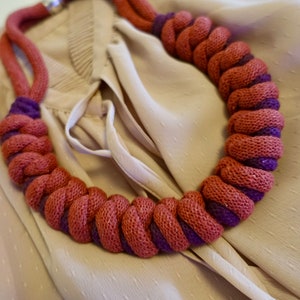 Può includere: Un primo piano di una collana spessa fatta a mano in tessuto intrecciato. La collana è una miscela di tessuto rosso scuro e viola, creando un look unico e testurizzato.