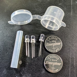 Op de afbeelding: Drie zilveren LED-lampjes, twee zilveren CR2025-batterijen met het label "Energizer" met "3 VOLTS" en "MADE IN JAPAN", een doorzichtige plastic container met een deksel en een doorzichtige plastic buis.