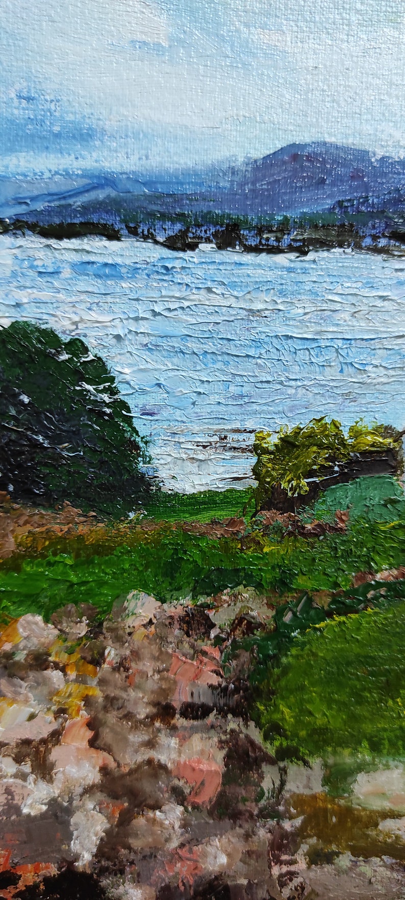 May include: A painting of a landscape with a lake in the background. The lake is a light blue color and the water is textured. There are green trees on either side of the lake. The foreground is a green grassy area with brown rocks.