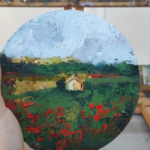 Op de afbeelding: Een rond schilderij op een houten ezel toont een landschap met een klein wit huis in de verte. Op de voorgrond is een veld met rode bloemen en groen gebladerte. De lucht is lichtblauw met witte wolken.
