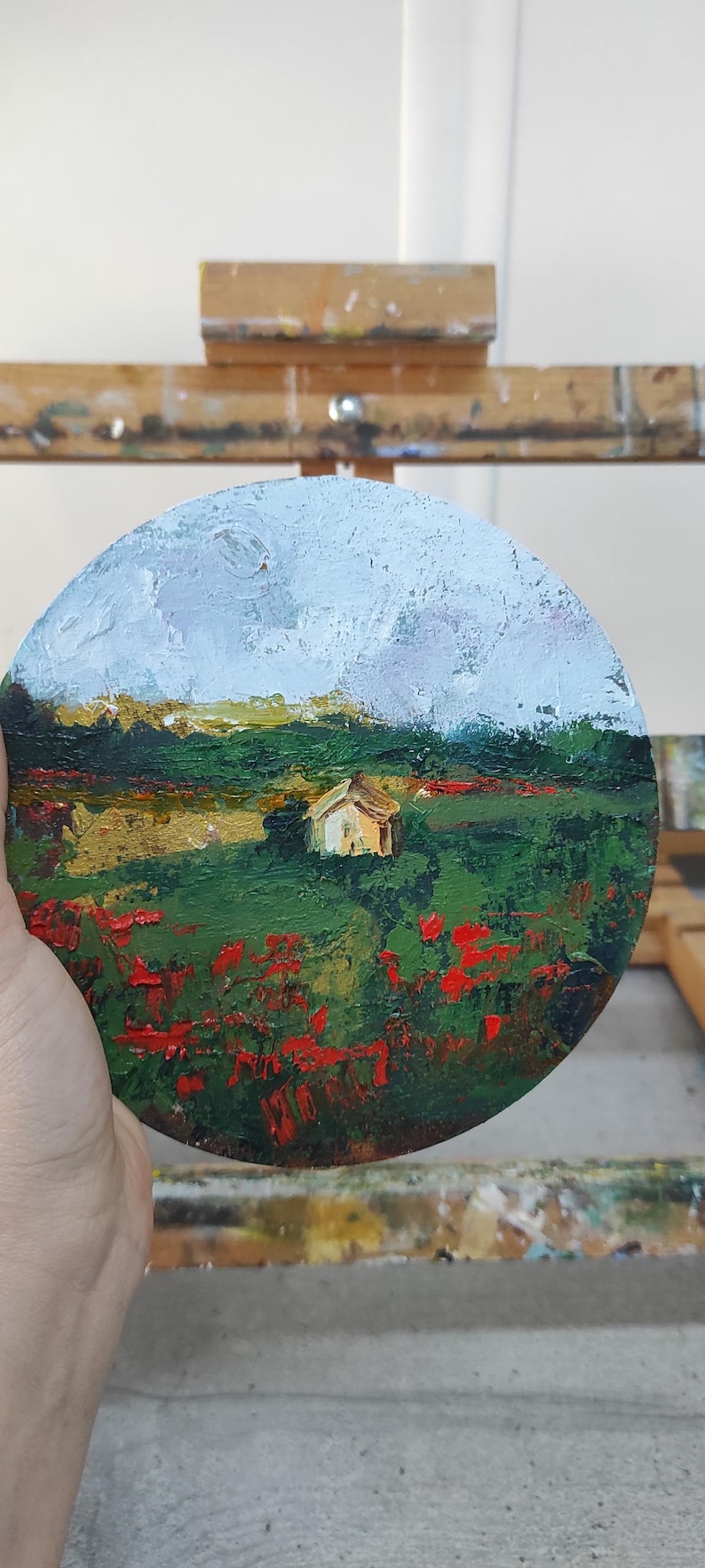 Op de afbeelding: Een rond schilderij op een houten ezel toont een landschap met een klein wit huis in de verte. Op de voorgrond is een veld met rode bloemen en groen gebladerte. De lucht is lichtblauw met witte wolken.