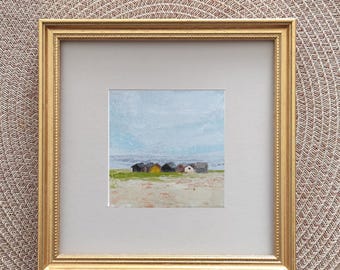 May include: A square painting in a gold frame featuring a landscape with a sandy beach, green grass, and a row of small beach huts under a light blue sky. The artwork is set against a light gray matting within the ornate gold-colored frame.