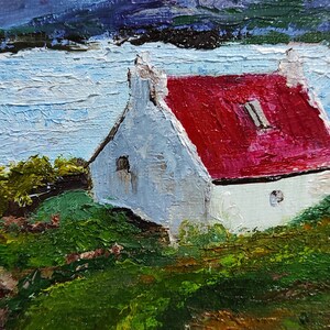 May include: A white cottage with a red roof sits on a grassy hill overlooking a body of water. The cottage has two windows and a chimney. The water is blue and the sky is light blue. In the background, there are mountains.