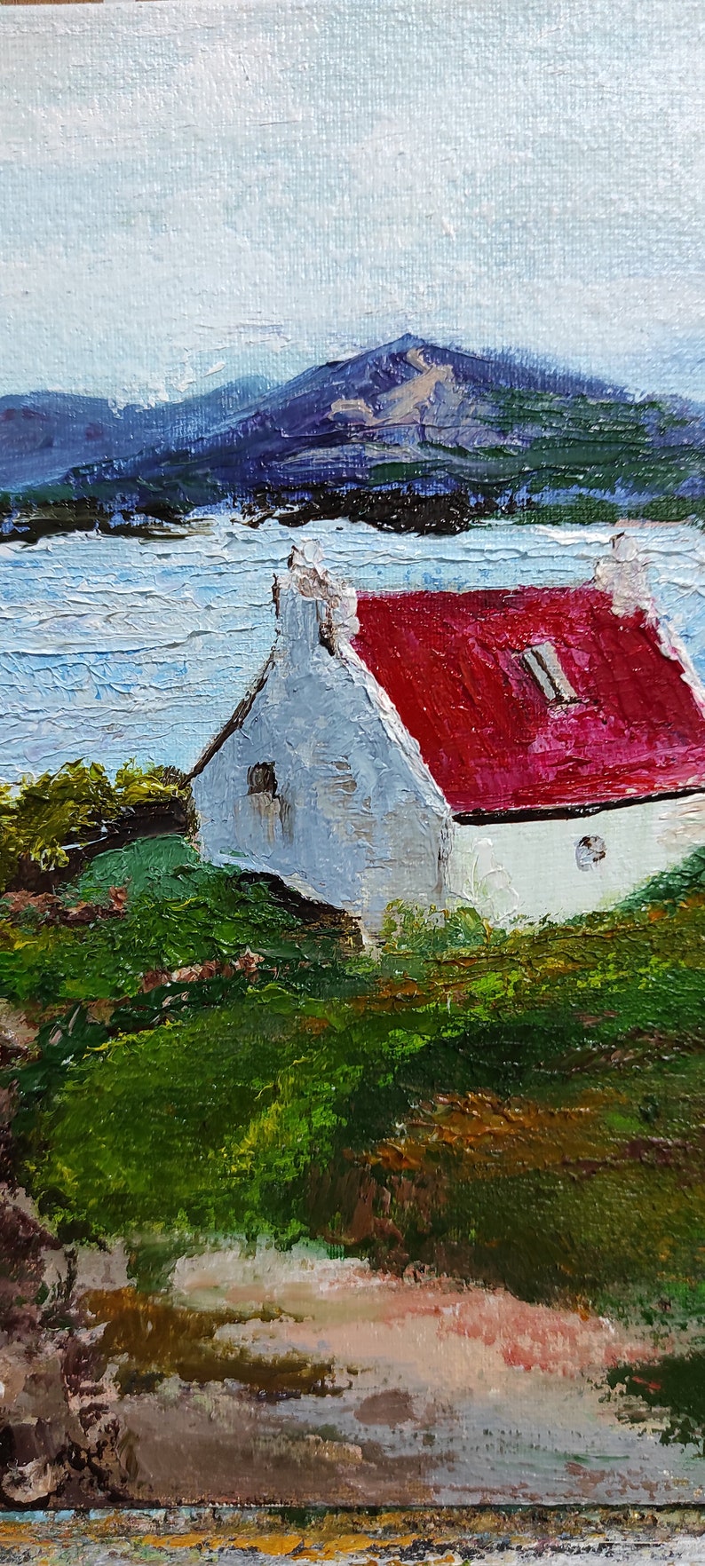 May include: A white cottage with a red roof sits on a grassy hill overlooking a body of water. The cottage has two windows and a chimney. The water is blue and the sky is light blue. In the background, there are mountains.