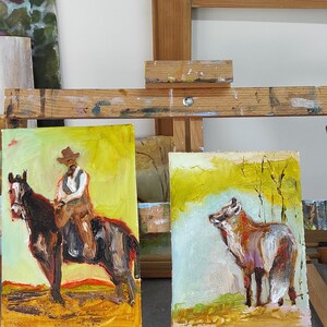 May include: Two oil paintings on canvas. One depicts a cowboy on a horse, the other a fox in a field. Both paintings have a yellow and brown color palette and are displayed on an easel.