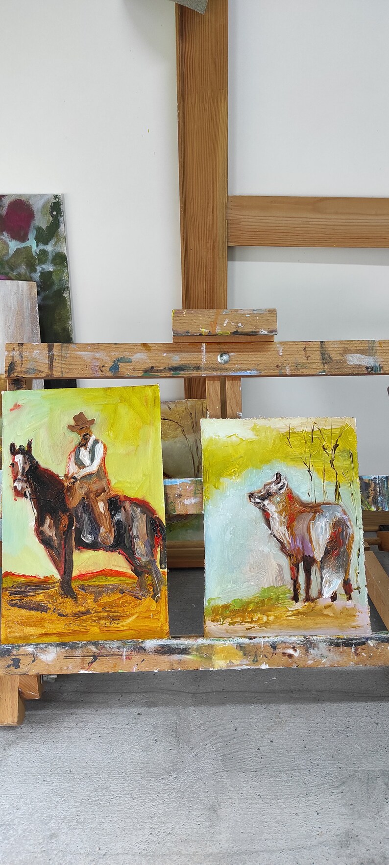 May include: Two oil paintings on canvas. One depicts a cowboy on a horse, the other a fox in a field. Both paintings have a yellow and brown color palette and are displayed on an easel.