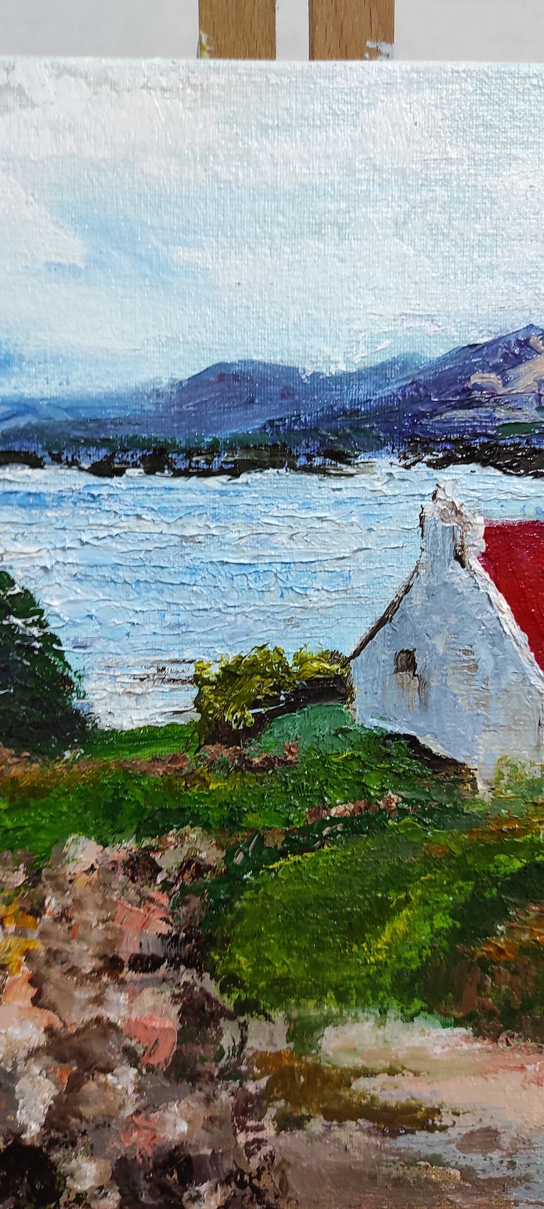 May include: A painting of a white cottage with a red roof, set against a backdrop of a blue lake and distant mountains. The cottage is surrounded by green grass and trees, and the sky is a light blue with white clouds.