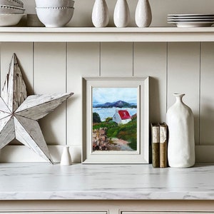 May include: A framed painting of a white cottage with a red roof on a green hill overlooking a body of water. The painting is on a white shelf with other decorative items, including a white vase, a white star, and a stack of books.
