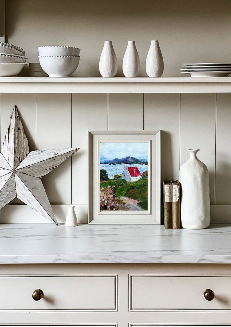 May include: A framed painting of a white cottage with a red roof on a green hill overlooking a body of water. The painting is on a white shelf with other decorative items, including a white vase, a white star, and a stack of books.