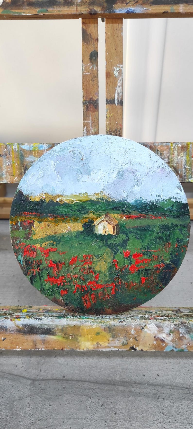 Op de afbeelding: Een rond canvas schilderij van een landschap met een klein huis in de verte. Op de voorgrond is een veld met rode bloemen en groen gras. De lucht is lichtblauw met witte wolken.