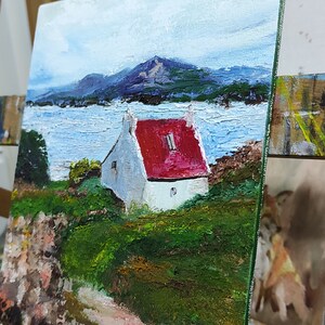 May include: A painting of a white house with a red roof, set against a backdrop of a blue lake and mountains. The house is surrounded by green grass and trees. The painting is on an easel.