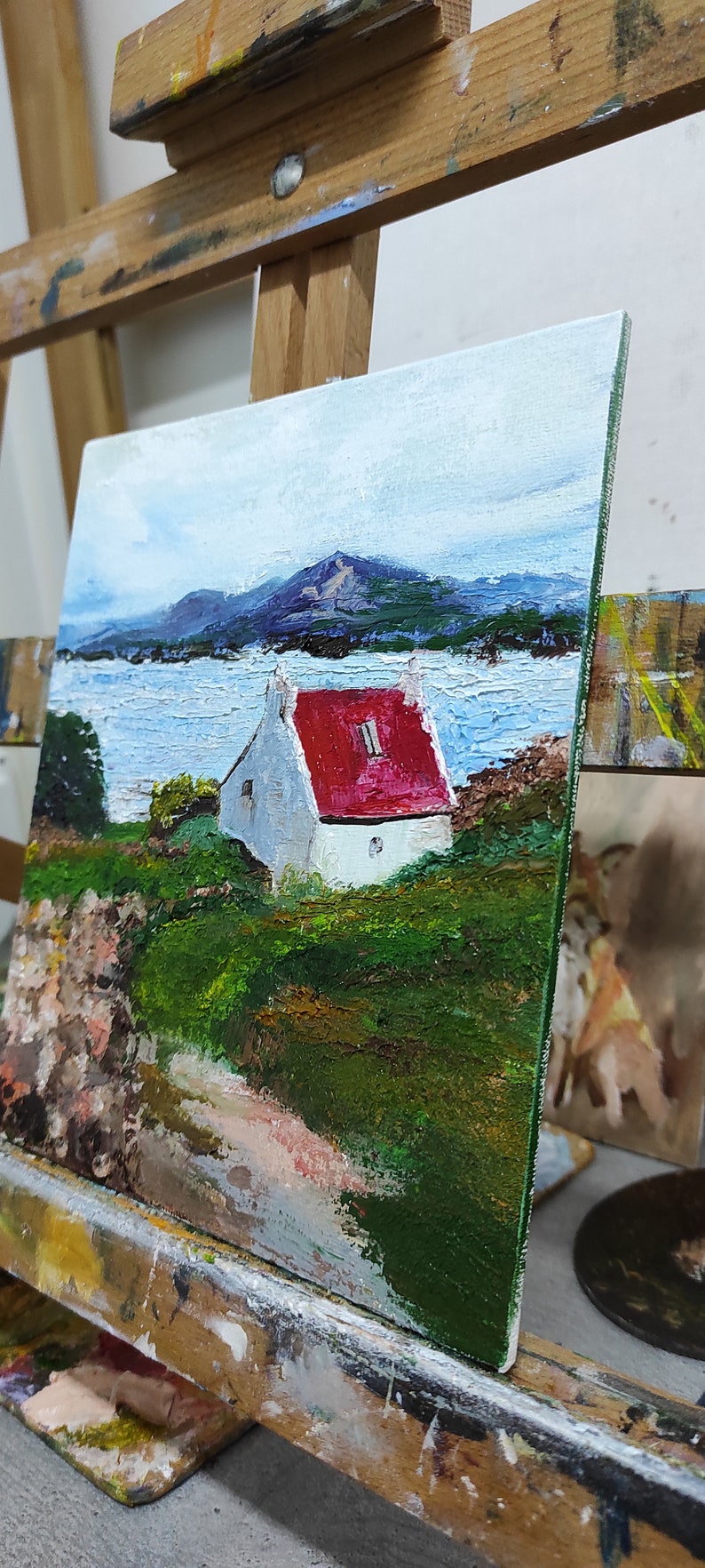 May include: A painting of a white house with a red roof, set against a backdrop of a blue lake and mountains. The house is surrounded by green grass and trees. The painting is on an easel.