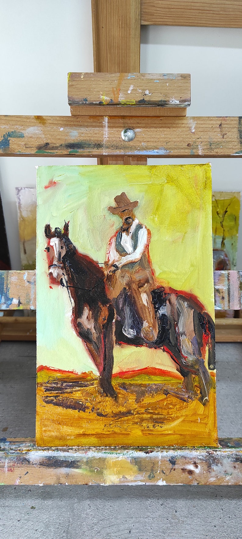 May include: Oil painting of a cowboy on horseback. The artwork features a cowboy in a brown hat and vest, riding a brown horse. The background is a blend of yellow and orange hues, suggesting a sunset or desert landscape.