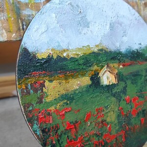 Op de afbeelding: Een rond canvas schilderij van een landschap met een klein wit huis in de verte. Op de voorgrond een veld met rode bloemen, en op de achtergrond een blauwe lucht met witte wolken.
