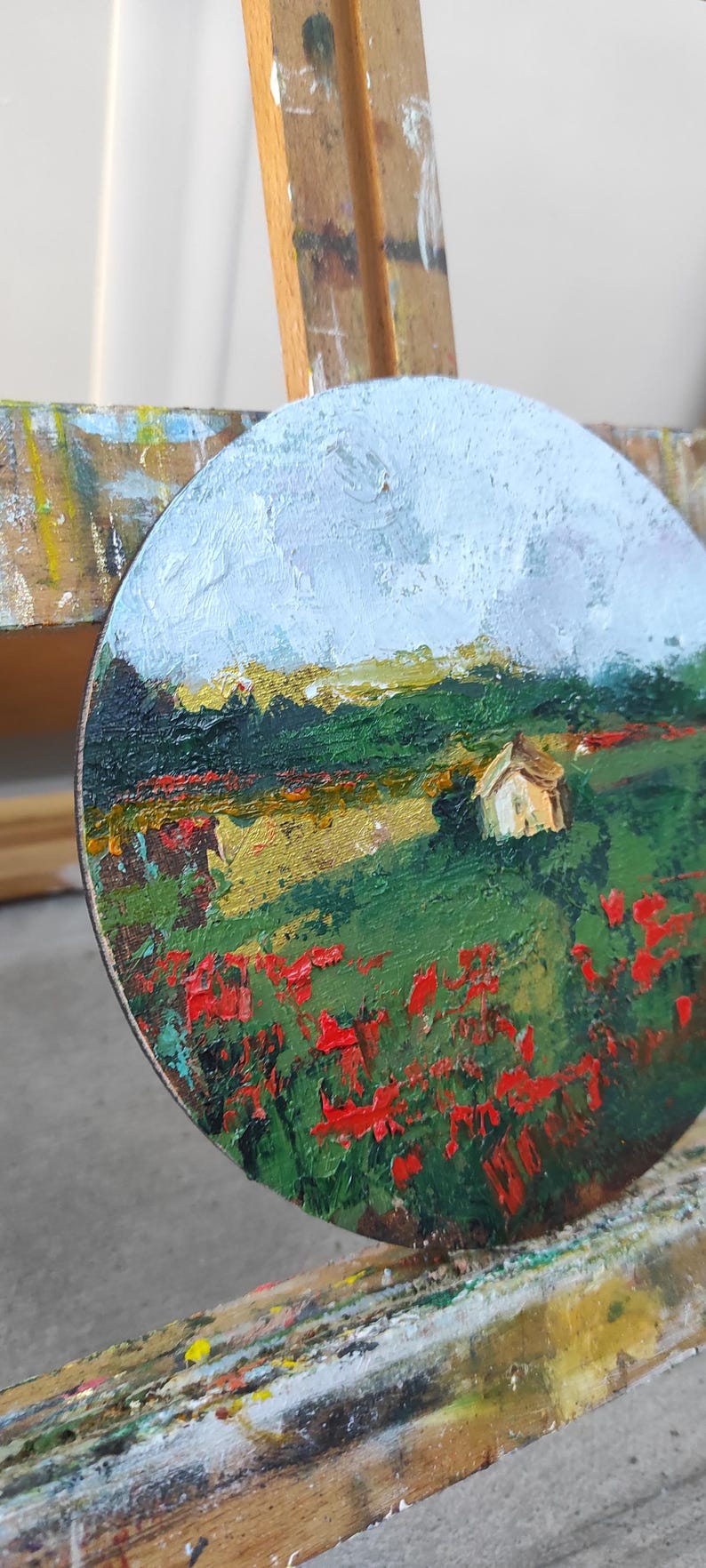 Op de afbeelding: Een rond canvas schilderij van een landschap met een klein wit huis in de verte. Op de voorgrond een veld met rode bloemen, en op de achtergrond een blauwe lucht met witte wolken.