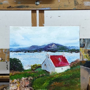 May include: A painting on an easel depicting a white cottage with a red roof, set against a backdrop of a blue lake and distant mountains. The painting is framed and has a textured brushstroke style.