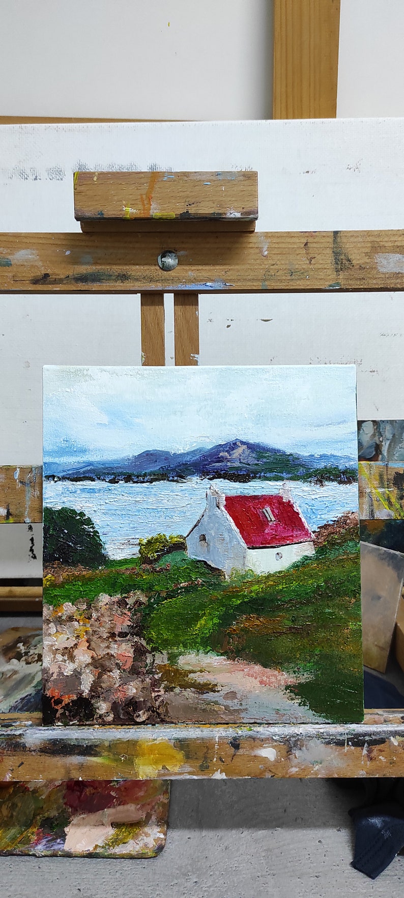 May include: A painting on an easel depicting a white cottage with a red roof, set against a backdrop of a blue lake and distant mountains. The painting is framed and has a textured brushstroke style.