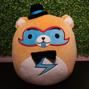 May include: A plush toy shaped like a bear wearing a black top hat with a blue band, a blue mask with red trim, a black bow tie, and a lightning bolt design on its chest. The bear is orange and white.