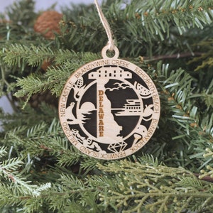 May include: A wooden ornament shaped like the state of Delaware with the word "Delaware" written vertically down the center. The ornament is black with the state outline and the word "Delaware" in natural wood color. The ornament is surrounded by a circle with the names of cities and towns in Delaware written around the edge.