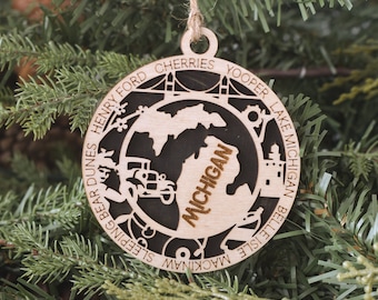 May include: A wooden Christmas ornament shaped like the state of Michigan with the word "Michigan" written inside. The ornament is decorated with various landmarks and locations around the state, including Mackinaw Island, Sleeping Bear Dunes, Henry Ford, Belle Isle, Yooper Lake Michigan, and Cherry Festival.