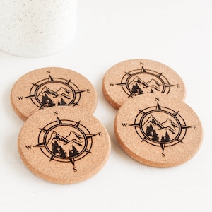 May include: Four round cork coasters with a compass design. Each coaster features a black compass rose with mountain and tree illustrations. The coasters are a natural tan color and are arranged on a white surface.