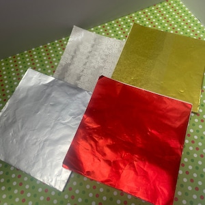 May include: An assortment of metallic wrapping paper sheets in silver, gold, and red. One sheet has a snowflake pattern. The sheets are arranged on a green surface with white and red polka dots, suggesting a holiday theme.