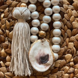 May include: A decorative beaded garland with large, off-white wooden beads, gold accents, and a natural oyster shell pendant. A beige tassel hangs from the side. The garland is displayed on a woven, textured surface.