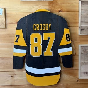 May include: A wooden wall art piece shaped like a hockey jersey, featuring a black base with yellow and white accents. The name CROSBY is printed in yellow above the number 87, with the number 87 on the sleeves in black and yellow.