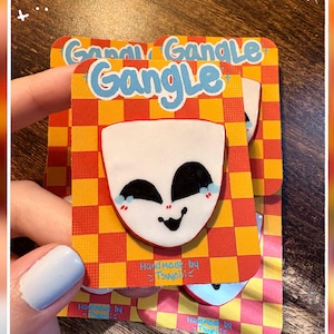 May include: A stack of three "Gangle" pins, each featuring a white face with black eyes and a smile, on a red and yellow checkered background. The pins are held by a hand with a light blue painted nail. Text on the card reads "Handmade by Tsuyoi<".