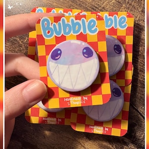 May include: A circular pin featuring a light purple creature with large dark eyes and a wide mouth filled with sharp white teeth. The pin is attached to a rectangular card with a yellow and orange checkerboard pattern and the text Bubble.