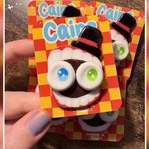 May include: A pin featuring a cartoon character with a black top hat, large white teeth, and mismatched blue and green eyes. The character is mounted on a rectangular backing card with a red and yellow checkerboard pattern and the text "Caine".