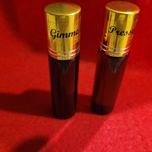 May include: Two dark brown glass bottles with gold-colored caps. The caps have the words "Gimme" and "Press" printed on them in black. The bottles are on a red surface.
