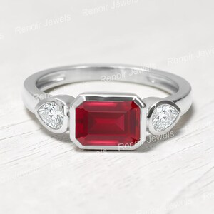 Könnte beinhalten: Ein silberner Ring mit einem rechteckigen, tiefroten Edelstein in der Mitte, flankiert von zwei birnenförmigen, klaren Edelsteinen. Der Ring hat ein glattes Band und eine Zargenfassung.