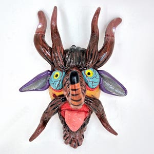 May include: A whimsical, handcrafted ceramic mask featuring a creature with brown horns, large blue eyes, and a red tongue. The mask has a textured surface and is painted with vibrant colors, including purple and yellow accents. The mask is designed to be hung on a wall.