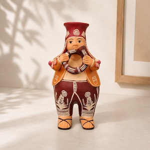 May include: A terracotta figurine of a person wearing a red hat, an orange vest, and dark red pants with white floral patterns. The figure holds a circular, striped instrument to their mouth and wears simple sandals on their feet.