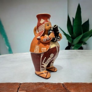 Może przedstawiać: Ceramiczna figurka osoby w tradycyjnym stroju, trzymającej czarny instrument. Figurka jest pomalowana w odcieniach pomarańczu, brązu i bieli, ze skomplikowanymi wzorami na ubraniach. Figurka ma pękaty korpus.