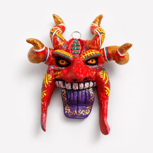 May include: A red, textured mask featuring four golden horns, yellow and white geometric patterns, yellow eyes with black pupils, a wide mouth with white teeth, and a large purple tongue hanging down. A small silver metal loop is attached at the top.