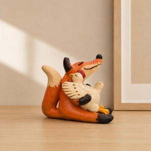 May include: A hand-painted figurine featuring an orange fox with black-tipped ears and paws, sitting and holding a small white chicken with a red comb. The fox has a curved tail and the chicken is positioned against the fox's chest in a resting pose.