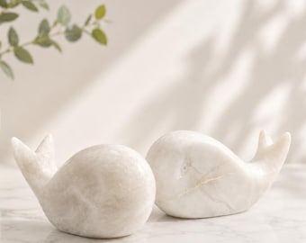 Può includere: Due statuette di balena in pietra bianca liscia e lucida. Ogni scultura presenta un corpo arrotondato, un dettaglio dell'occhio e una pinna caudale curva verso l'alto. Le venature naturali della pietra sono visibili su entrambe le balene.