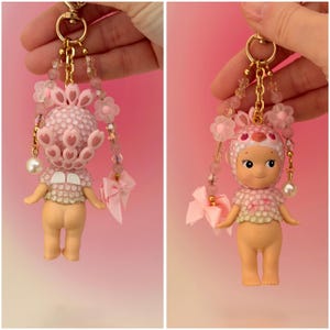 Puede incluir: Un llavero de mu&ntilde;eca rosa con una cadena dorada, adornado con cuentas rosas, flores y un lazo. La mu&ntilde;eca lleva un atuendo rosa y un tocado. Una perla cuelga de la cadena. Es un art&iacute;culo de colecci&oacute;n.