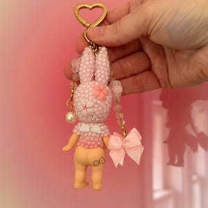 Puede incluir: Un llavero de conejito rosa con un cierre dorado en forma de coraz&oacute;n. El conejito est&aacute; cubierto de peque&ntilde;as cuentas brillantes y tiene un lazo rosa y una flor en la cabeza. El llavero se sostiene sobre un fondo rosa.