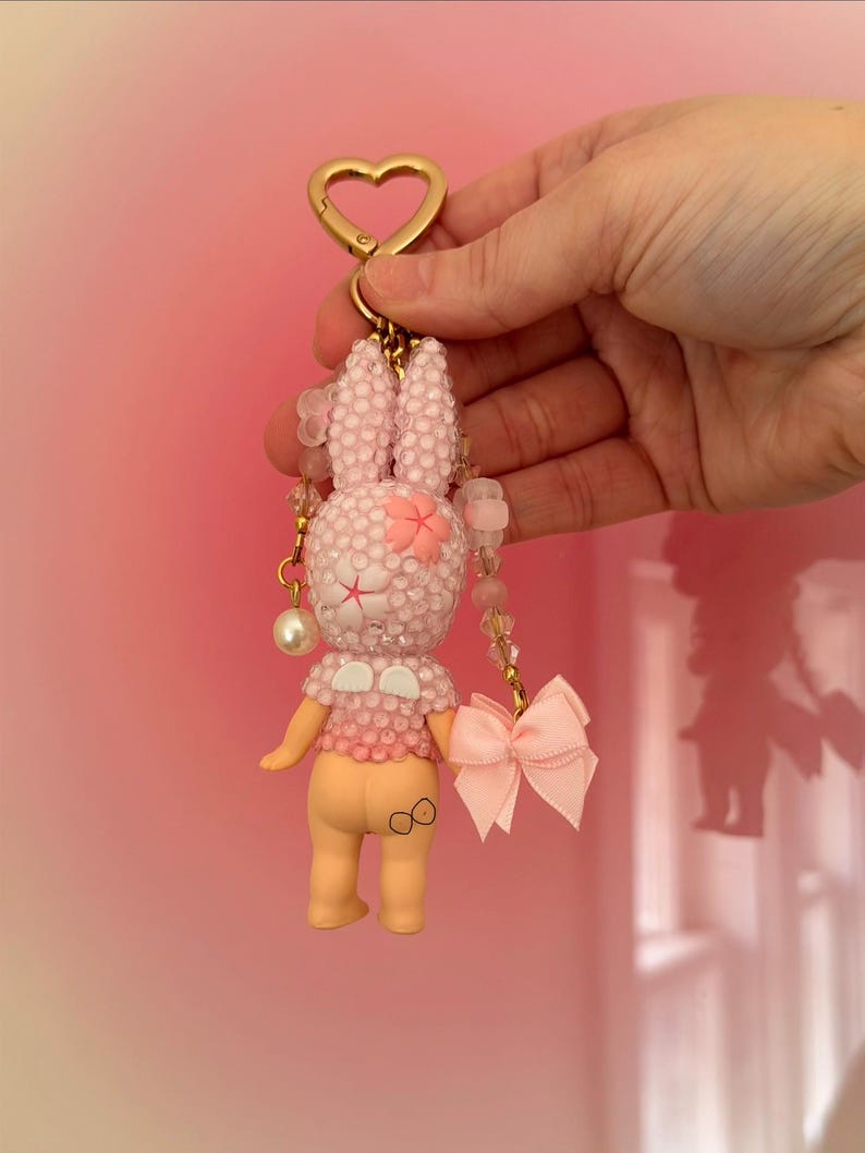 Puede incluir: Un llavero de conejito rosa con un cierre dorado en forma de coraz&oacute;n. El conejito est&aacute; cubierto de peque&ntilde;as cuentas brillantes y tiene un lazo rosa y una flor en la cabeza. El llavero se sostiene sobre un fondo rosa.