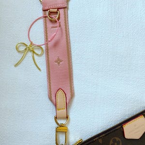 K&ouml;nnte beinhalten: Ein rosa und braunes Schulterband mit goldenen Beschl&auml;gen und einem kleinen goldenen Schleifenanh&auml;nger. Das Band ist an einer braunen Ledertasche mit dem Louis Vuitton Logo befestigt.