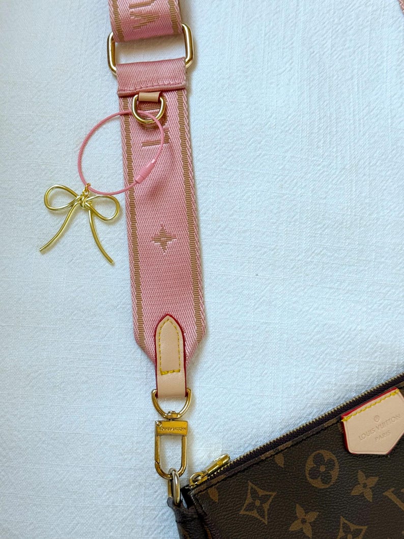 K&ouml;nnte beinhalten: Ein rosa und braunes Schulterband mit goldenen Beschl&auml;gen und einem kleinen goldenen Schleifenanh&auml;nger. Das Band ist an einer braunen Ledertasche mit dem Louis Vuitton Logo befestigt.