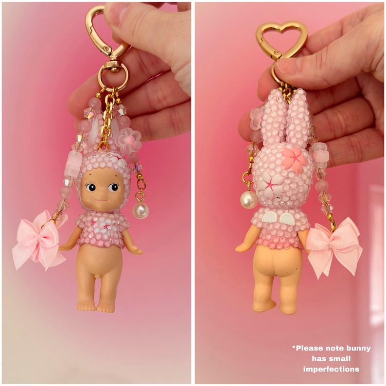 Puede incluir: Un llavero rosa y blanco con cuentas que presenta una peque&ntilde;a mu&ntilde;eca con una parte superior decorada con flores y un lazo rosa. La mu&ntilde;eca tiene un tono de piel claro y est&aacute; unida a un cierre dorado con dise&ntilde;o de coraz&oacute;n.