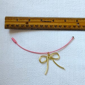 K&ouml;nnte beinhalten: Eine rosafarbene Schnurkette mit einem Anh&auml;nger aus goldenem Draht in Form einer Schleife.