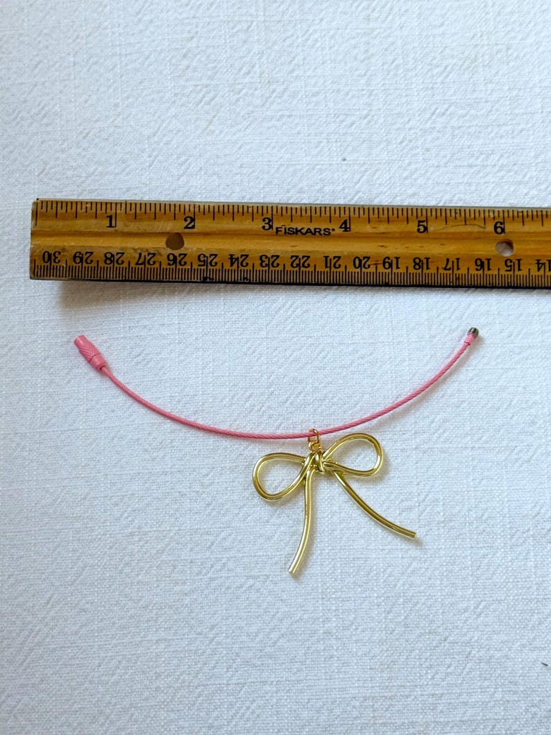 K&ouml;nnte beinhalten: Eine rosafarbene Schnurkette mit einem Anh&auml;nger aus goldenem Draht in Form einer Schleife.