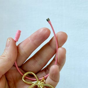 K&ouml;nnte beinhalten: Eine rosa Schnur mit einem goldenen Schleifenanh&auml;nger. Die Schnur hat an einem Ende eine Metallkappe und am anderen Ende eine rosa Plastikkappe.