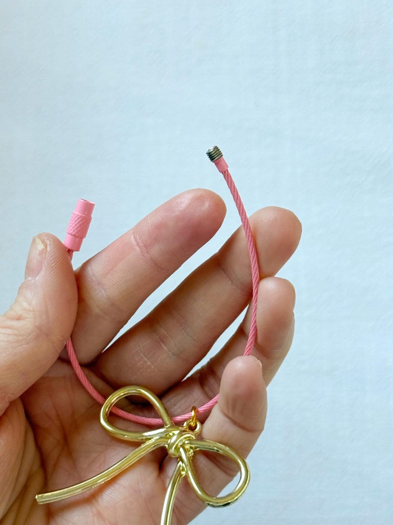 K&ouml;nnte beinhalten: Eine rosa Schnur mit einem goldenen Schleifenanh&auml;nger. Die Schnur hat an einem Ende eine Metallkappe und am anderen Ende eine rosa Plastikkappe.