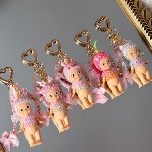 Puede incluir: Cinco llaveros con peque&ntilde;as figuras de mu&ntilde;ecas. Cada mu&ntilde;eca lleva un atuendo de cuentas rosas y blancas, alas y cierres dorados en forma de coraz&oacute;n. Los tocados var&iacute;an, incluyendo orejas de conejo y dise&ntilde;os florales. Cintas y cuentas rosas.